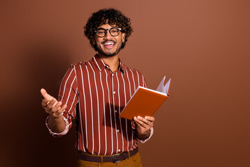 Portrait of nice young man hold book speak wear striped shirt isolated on brown color background