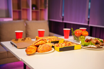 An indoor office party scene with no people around, featuring a desk filled with food pizza, bread, salad, chicken grill, French fries, and beerset against a night light with RGB lighting.