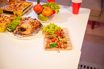 An indoor office party scene with no people around, featuring a desk filled with food pizza, bread, salad, chicken grill, French fries, and beerset against a night light with RGB lighting.