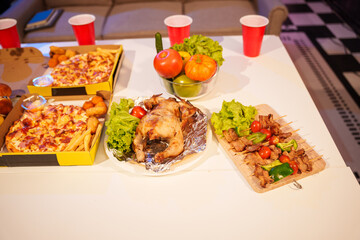 An indoor office party scene with no people around, featuring a desk filled with food pizza, bread, salad, chicken grill, French fries, and beerset against a night light with RGB lighting.