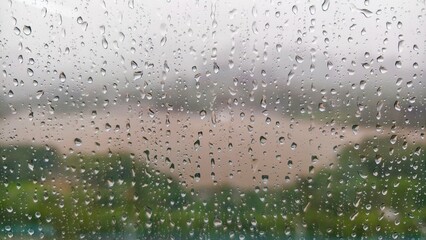 It's raining heavily outside the window, raindrops are falling on the window, and you can vaguely see the green trees and the yellow lake in the distance.
