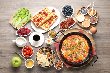 Tasty shakshouka in frying pan and other meals served for breakfast on wooden table, flat lay