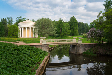 Obraz premium View of the Temple of Friendship on the banks of the Slavyanka River against the background of the Cast-iron bridge in Pavlovsky Park on a sunny summer day, Pavlovsk, St. Petersburg, Russia