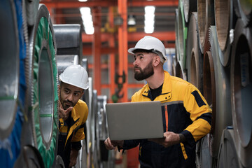 Metalwork manufacturing, warehouse for galvanized or metal sheet. Team of male engineer factory worker using laptop computer inspecting quality rolls of galvanized or metal sheet in aluminum warehouse