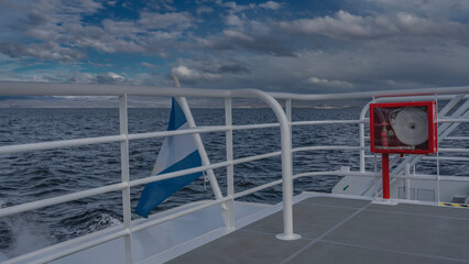 The national flag of Argentina is mounted on the ship's railing. A blue-and-white cloth flutters at the stern. Ripples on the ocean surface. Clouds in the blue sky. The Beagle Channel.Tierra del Fuego