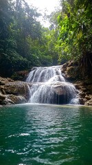 Fototapeta premium A cascading waterfall flows over rocks into a still, teal pool in a lush green jungle.