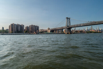 Naklejka premium This breathtaking view of the Manhattan Bridge features the iconic New York City skyline in the background. The clear blue skies reflect on the calm waters below, highlighting the city’s impressive ar