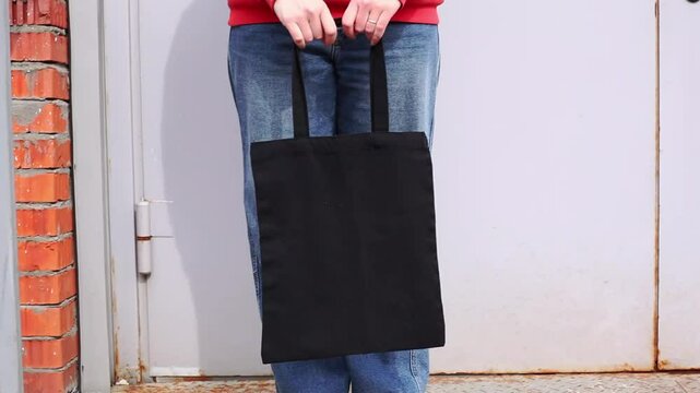 Woman in red hoodie holding tote canvas blank eco bag on street wall background. Female consumer hold black textile shopper. template or place for your design, text, logo
