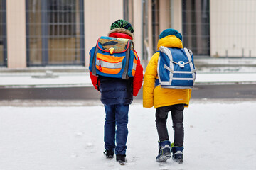 Two little kids boys of elementary class walking to school during snowfall. Happy children having fun and playing with first snow. Siblings and best friends with backpack in colorful winter clothes.