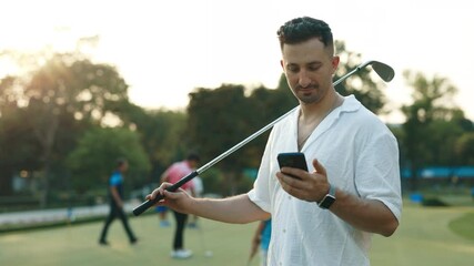 Portrait of bearded man with golf club looking at cellphone screen on sports ground. Male player reads news about competition winners in social media. Positive golfer bets online - Powered by Adobe