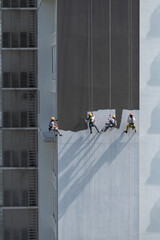 Four workers wearing colourful helmets hanging on ropes at height painting a tall building on a very sunny day.