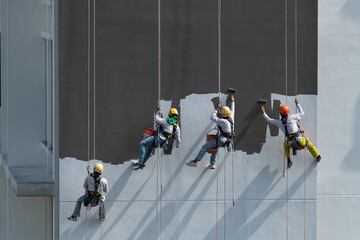 Four workers wearing colourful helmets hanging on ropes at height painting a tall building on a very sunny day.