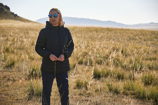 Woman in puffer jacket standing in grassy field, holding plant stalk.