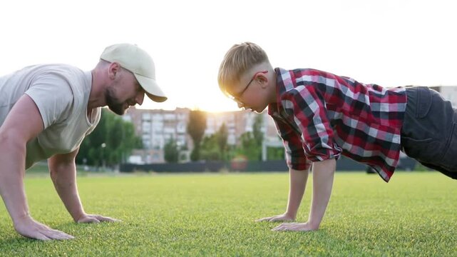 Young muscular father doing push-ups with his little son. Active happy family having fun on green grass. Fatherhood concept.