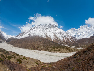 A panoramic view of a snow-capped mountain range. The sky is a clear blue, with a few puffy white...
