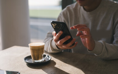 checking social media with smartphone drinking coffee in cafe against beautiful sunlight, having a relaxing moment