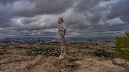 Naklejka premium A man stands on the edge of a cliff, looking into the distance. Hands on the belt. Profile view. In the distance and below is a panorama of Madagascar: fields, red-soil roads, rural houses the trees