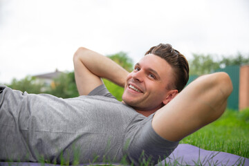 Man lies on a mat outdoors, smiling as he performs an abdominal exercise with his hands behind his head. The image captures a moment of fitness and joy, highlighting a positive and active lifestyle.