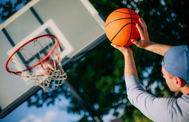 A close-up shot of a person in a blue cap aiming a basketball at an outdoor hoop. The vibrant orange ball contrasts with the blurred green foliage and blue sky in the background.