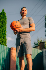 Fototapeta premium A man in a backward cap stands confidently, holding a basketball in both hands while looking off into the distance. The low-angle shot, warm sunlight, and clear sky enhance the strong image.