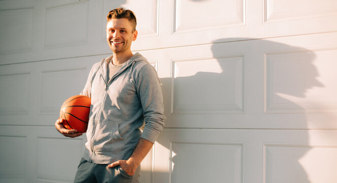 A smiling handsome man in a gray hoodie holds a basketball while standing against a sunlit garage door, casting a large shadow. The warm light and casual stance create a relaxed, sporty atmosphere.
