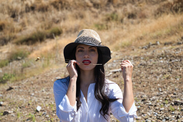 Portrait of a beautiful young brunette latin woman with red lips, dressed in a white shirt and straw hat, with her teeth killing a branch of dry grass. In the background there are trees and vegetation