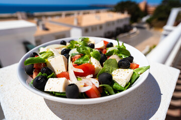 Fresh greek style salad with feta cheese in white bowl standing against backdrop of white houses and ocean and blue sky. Eating outdoors.