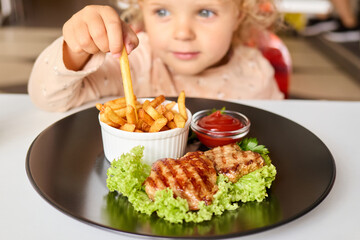 Little girl with curly blond hair sitting at the table in cafe enjoying grilled meat with potato free and sauce for lunch having fast food diner eating junk food with joy