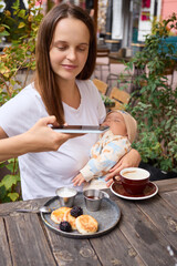 Smiling woman with newborn baby taking photo of delicious cheese pancakes with fresh blackberries on plate during morning breakfast making content for her private vlog photographing her breakfast