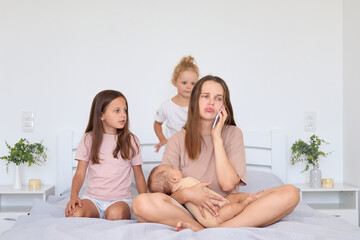 Upset mother holding newborn baby in hands sitting in bedroom with older children while talking on smartphone managing family and business at home having troubles expressing sadness