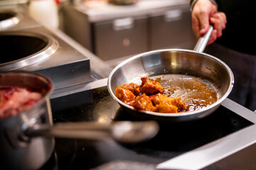 Close-up of a stainless steel frying pan on a stove with sizzling food, possibly meat in sauce, being cooked. The cook’s hands are visible, suggesting the action of cooking