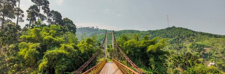 Obraz premium A suspension bridge stretches over lush green forested hills under a clear sky, representing adventure travel and eco-tourism