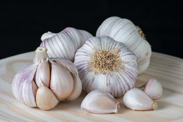 Fresh purple garlic heads and cloves on a rustic wooden plate with black background.