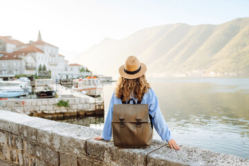 A cheerful woman poses by the water in a charming coastal town, wearing a hat and a light blue blouse, surrounded by serene mountains and boats. Europe travel.