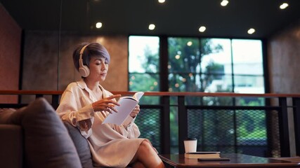 A mature woman listens to music through her headphones while reading a book in a cozy cafe. She sits comfortably with a coffee cup beside her, enjoying the peaceful ambiance