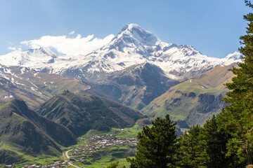 Gergeti village under the mountains, Gergeti Trinity Church on the hill and snow-capped Mkinvartsveri Kazbek. Blue sky with clouds