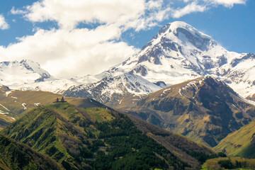 Fototapeta premium Gergeti Trinity Church on the hill and snow capped mountain Mkinvartsveri Kazbek. Blue sky with clouds