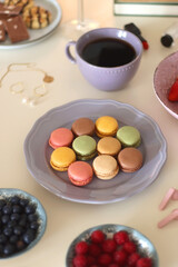 Plate of pastel macarons, cookies and chocolate, cup of tea of coffee, glass of bubble water, various berries, books and accessories on the table. Selective focus, pastel colors.
