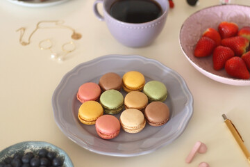 Plate of pastel macarons, cookies and chocolate, cup of tea of coffee, glass of bubble water, various berries, books and accessories on the table. Selective focus, pastel colors.