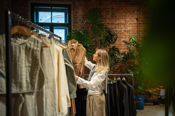 Interested woman stands in fashion store, carefully chooses clothes to buy. Pleased female looks at stylish dress while shopping, holds hanger with item, tries on likes, examining before make decision