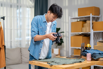 Young Asian man, distributor, online shop owner, small business owner, standing in warehouse, using camera to take pictures of products for sale.