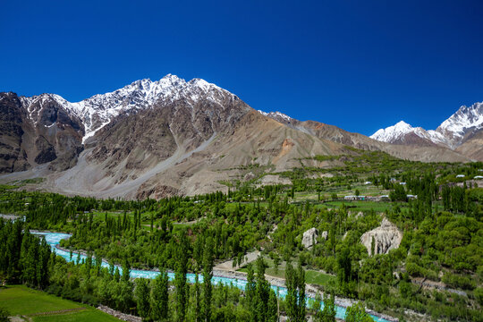 View of majestic snowcapped mountains and a tranquil river in a picturesque valley surrounded by lush greenery, Chitral, Pakistan.