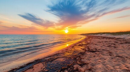 Obraz premium A serene sunset over a sandy beach with gentle waves and vibrant clouds reflecting golden hues in the sky.