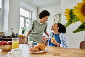 Two women share laughter and drinks while enjoying breakfast together at home.