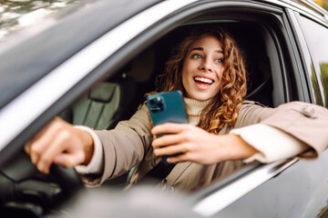 A young woman with curly hair enjoys driving by smiling and using her smartphone, capturing a...