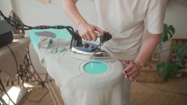 Camera focus on person's hands using pressing iron. Carefully removing any wrinkles on clothing. Getting fabric piece ready for next step. Textile laying on ironing board for stability.