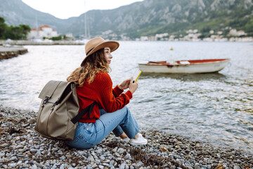 Young woman sitting on a wooden dock by the water, enjoying a sunny day while using her smartphone in a picturesque location. Woman shopping online, viewing social media or chatting in mobile app.