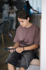 Boy plugging in his phone to a wired charger. Three females cooking in the kitchen on background