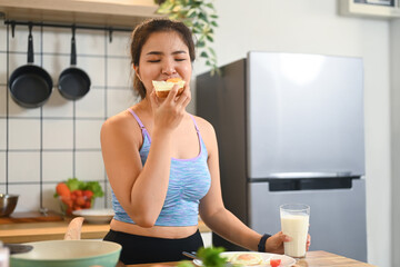 Young woman eating a sandwich and drinking milk, enjoying her meal in a cozy kitchen