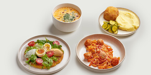 A variety of delicious and appetizing dishes are arranged on a white surface. The dishes include a bowl of creamy soup, a plate of meatballs and pasta, a plate of mashed potatoes